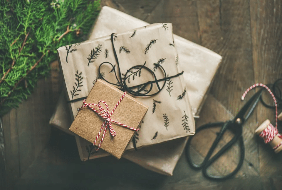 3 grijze en bruine geschenkverpakkingen, omringd door cadeaulinten, geplaatst op een bruine houten vloer met een beitel en een sparrentak op de achtergrond