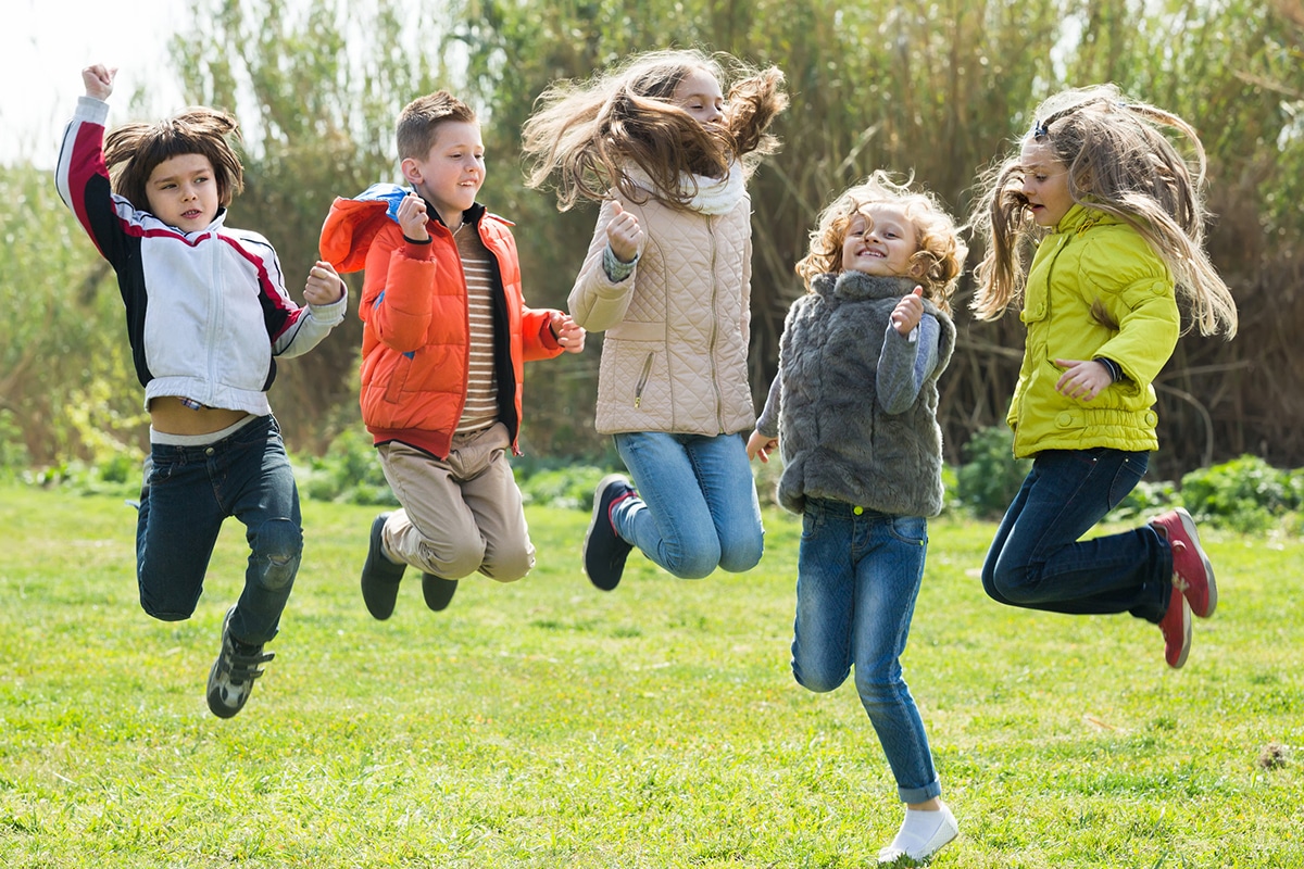 Een groep blije kinderen springt op het gras in de buitenlucht. Er zijn 2 jongeren en 3 meisjes. Ze zijn op het winkelcentrum