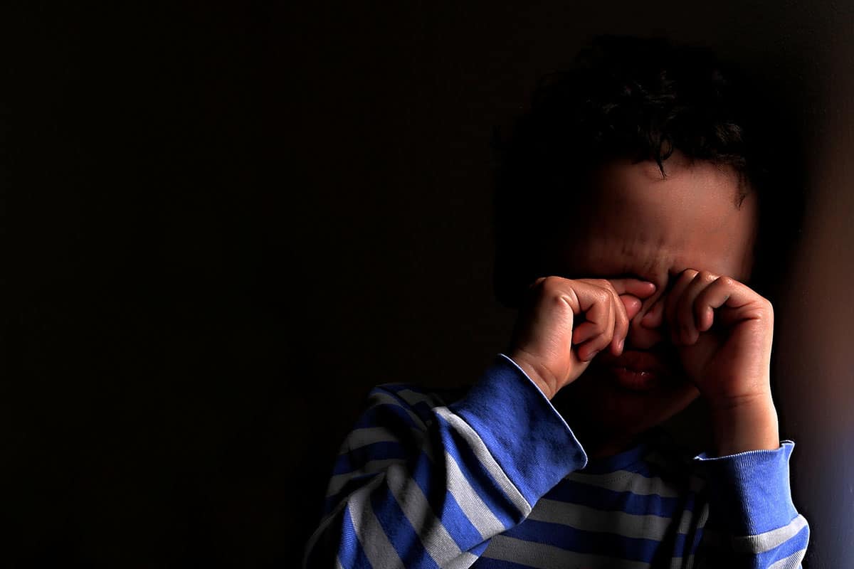 Een kleine jongen van gemengd ras in een blauw-wit gestreepte trui. De jongen houdt zijn ogen vast omdat hij bang is in het donker.