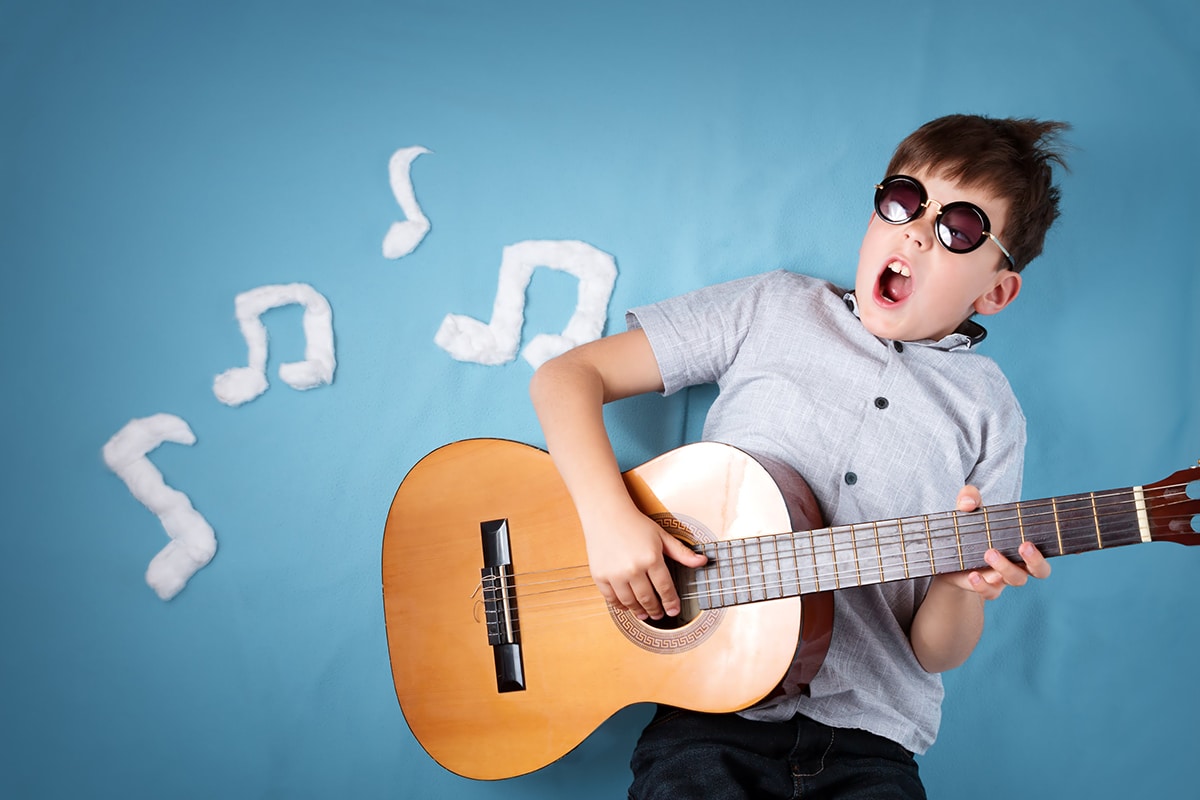 Een jongen speelt gitaar tegen een blauwe achtergrond. De jongen draagt een donkere bril, een grijs shirt met korte mouwen en een bruine houten gitaar. Er zijn witte muzieknoten op de achtergrond