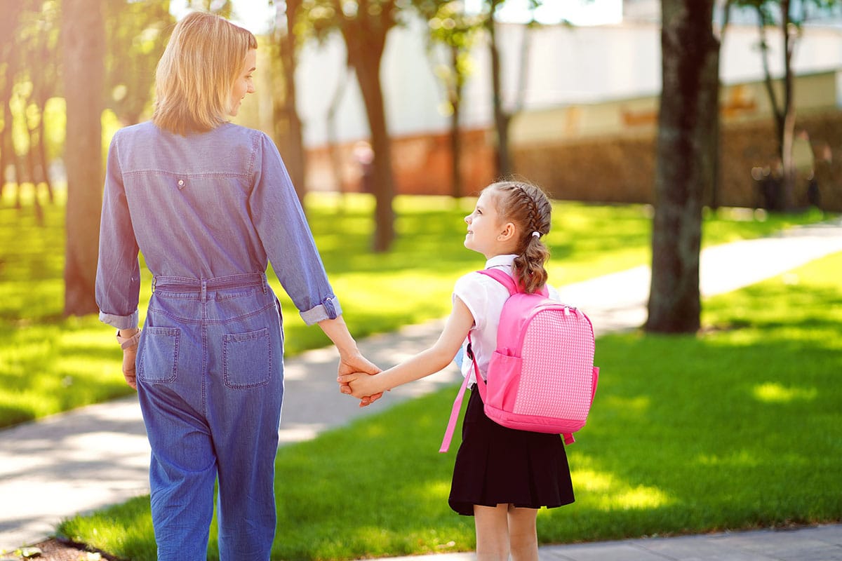 Een moeder neemt haar kind mee naar een park. Het is zonnig, de moeder is gekleed in een blauw spijkerbroek, het kleine meisje draagt een roze kinderrugzak