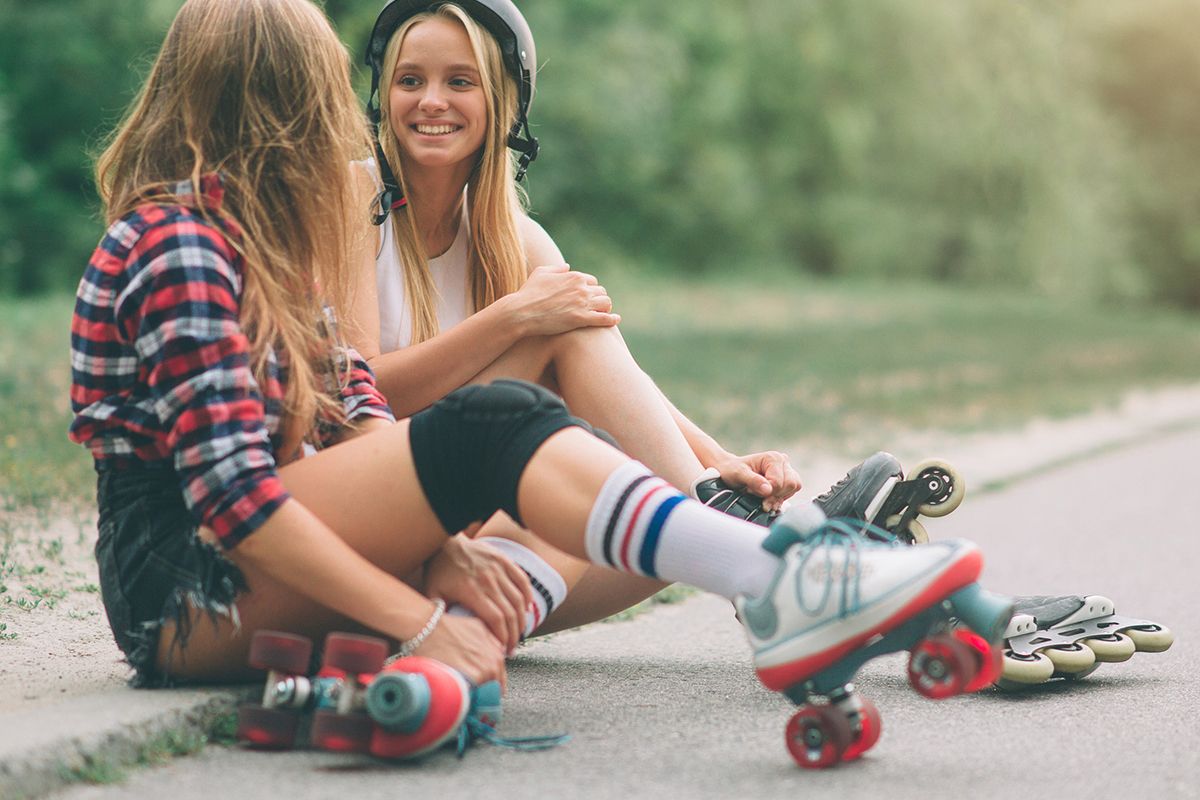 Twee jonge meisjes zitten aan de kant van de weg. De ene draagt rolschaatsen, de andere rolschaatsen en een helm.