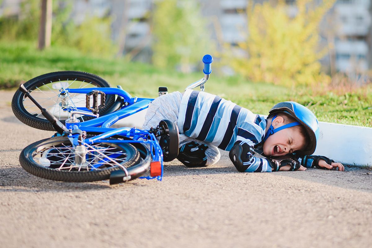 Een jong kind dat van zijn blauwe fiets is gevallen. Hij huilt en draagt een fietshelm, kniebeschermers en zwarte beschermende handschoenen