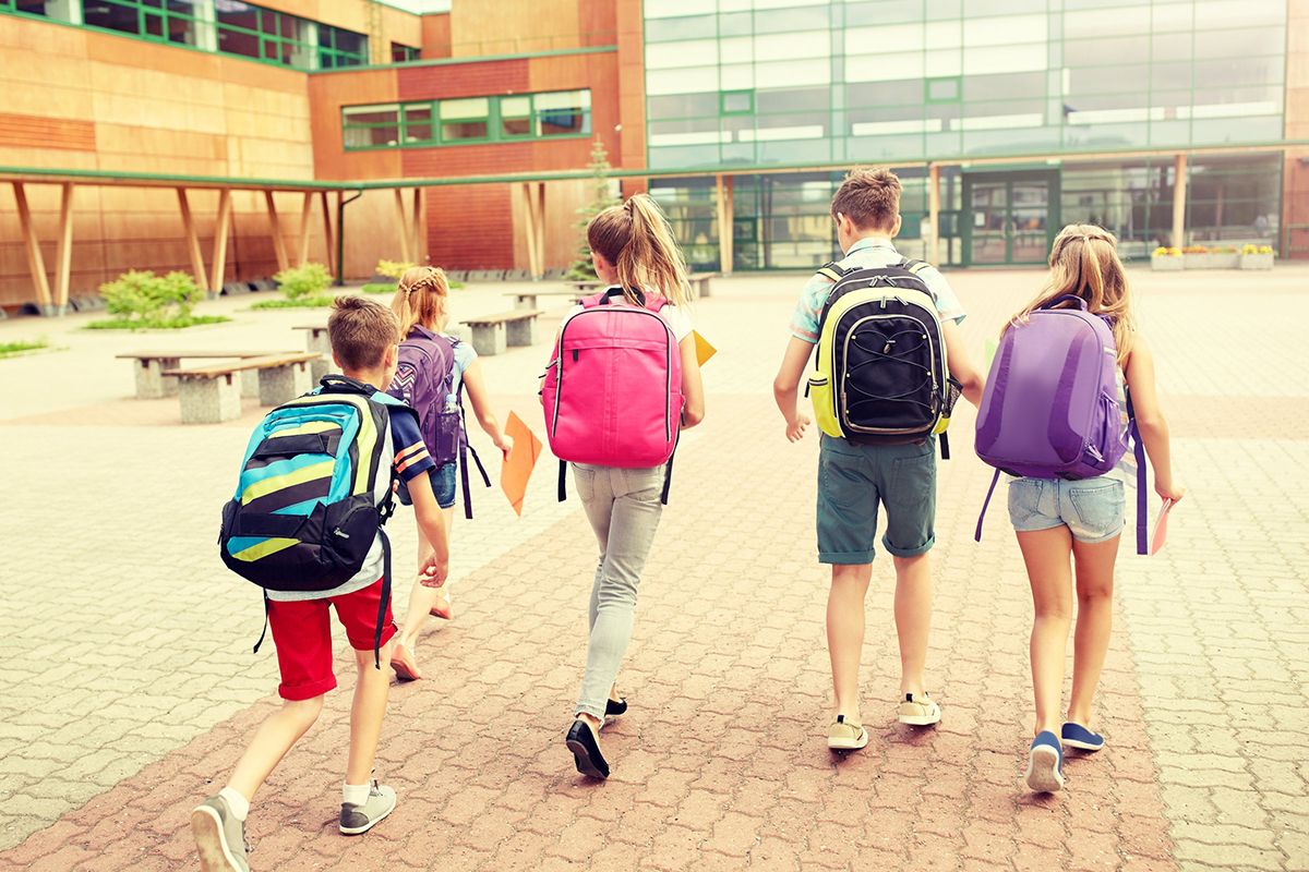 Een groep van 5 kinderen met de rug naar school. Elk kind draagt een gekleurde schooltas of rugzak