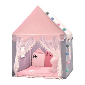 Een kindertipi in de vorm van een roze huisje, met een open voorkant en twee gordijnen die aan de zijkanten hangen. Er zitten kleine raampjes aan de zijkanten. Binnenin ligt een roze gewatteerde vloer en roze kussens.